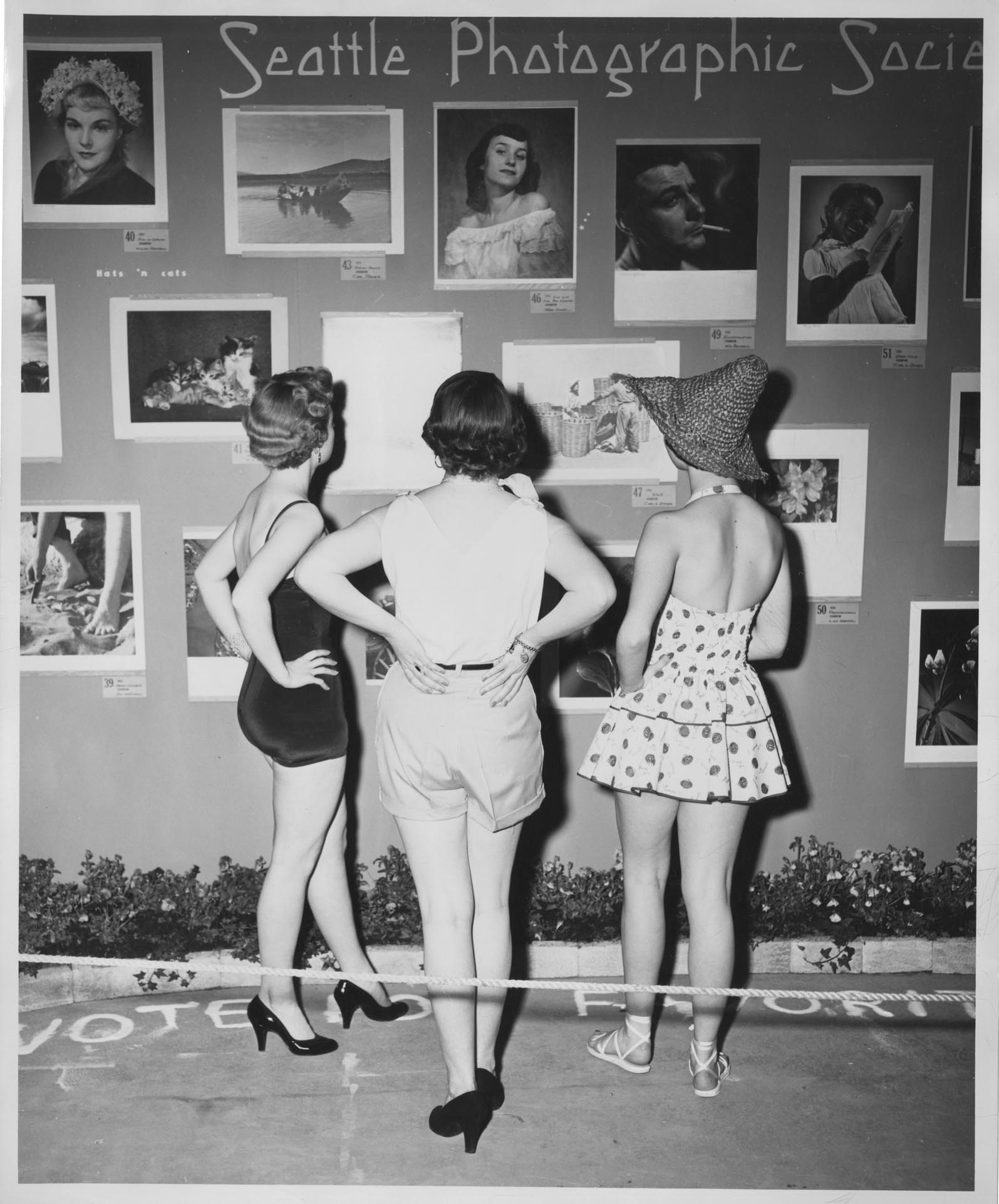 Image of Three women looking at photographs on exhibit, Seattle, circa 1950