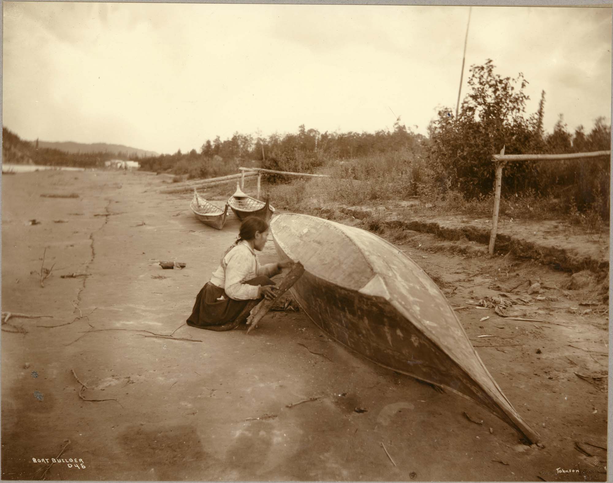 Image of Native Alaskan woman working on a canoe by a river, circa 1909