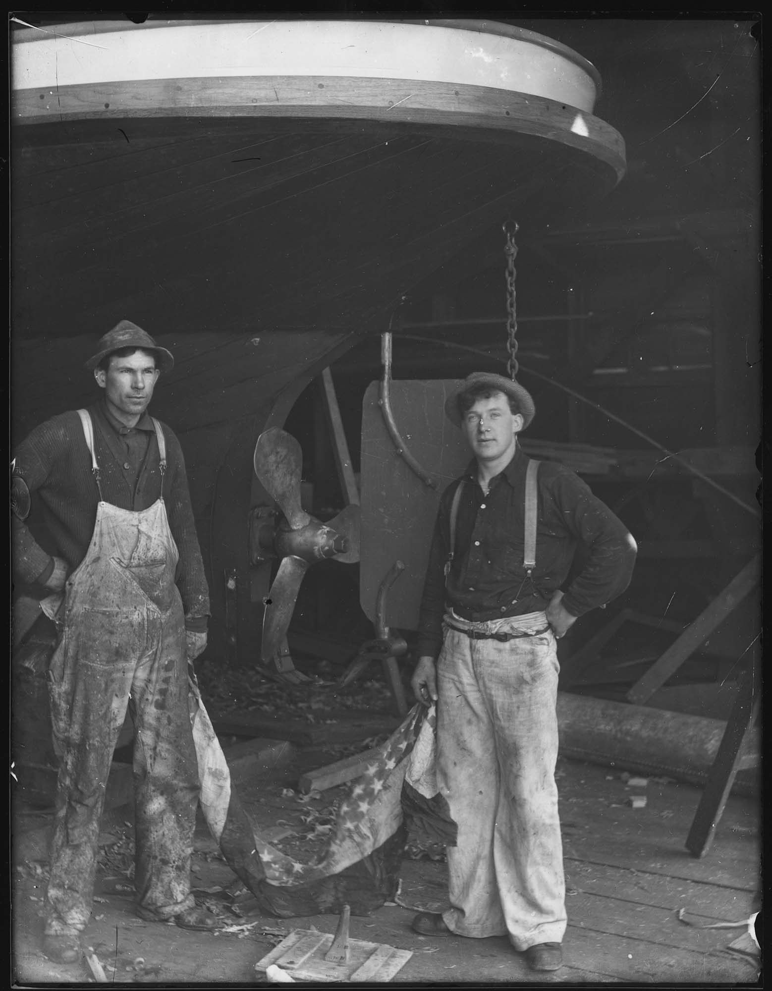 Image of Two men in front of ship hull holding flag, probably in Seattle, circa 1910