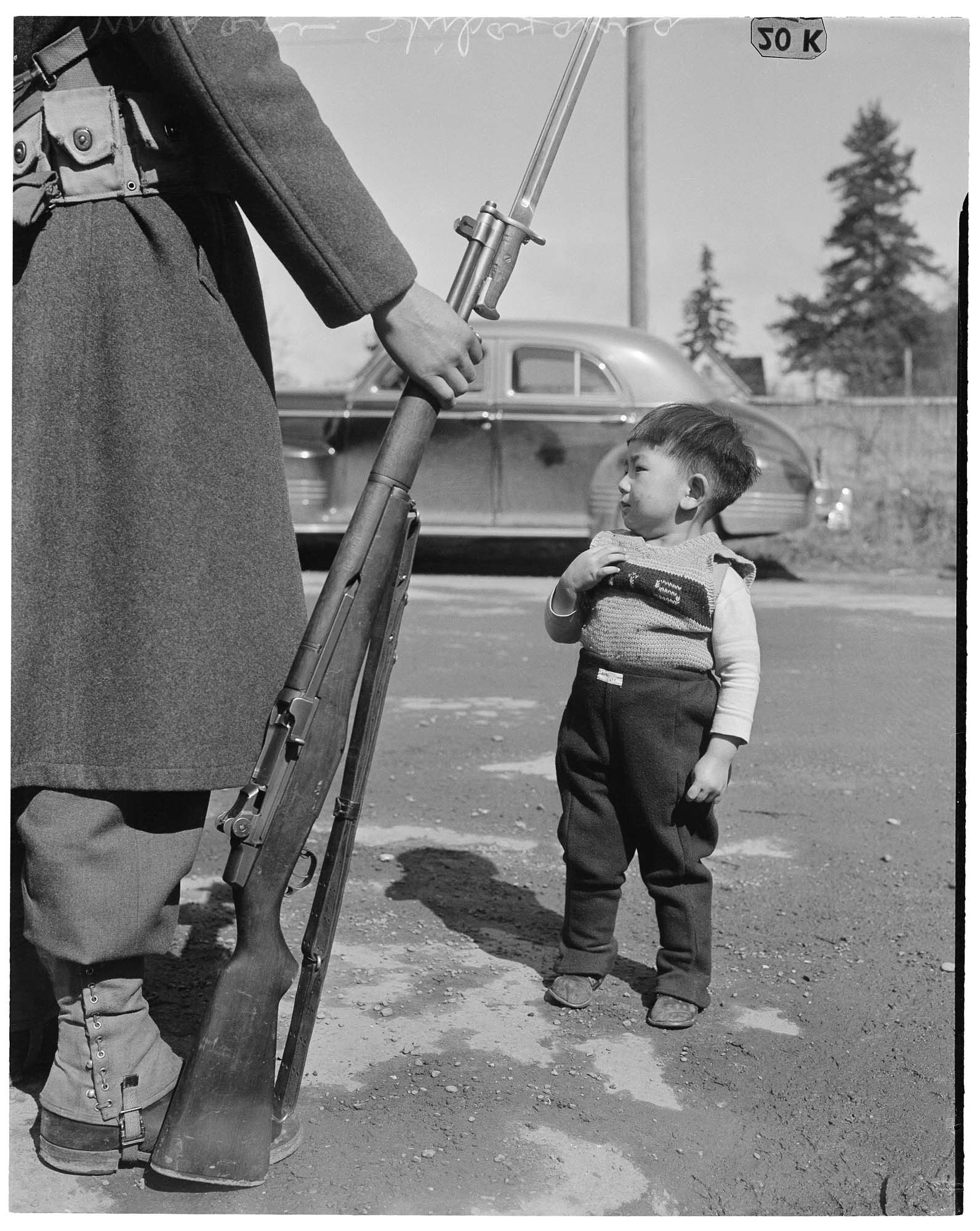 Image of Japanese American boy standing near a soldier with a rifle, possibly in Seattle, 1942